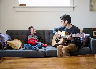 A cute boy with Downs Syndrome having fun playing guitar with his dad on the couch at home. he is holding a red ukulele and his dad is strumming an acoustic guitar and singing.